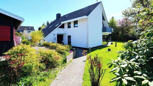Et moderne hus med to etager, skrå tag, belagt indkørsel og en velplejet have med forskellige planter og blomster.