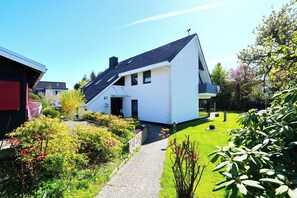 Una casa moderna de dos plantas con techo inclinado, entrada pavimentada y un jardín cuidado con diversas plantas y flores.