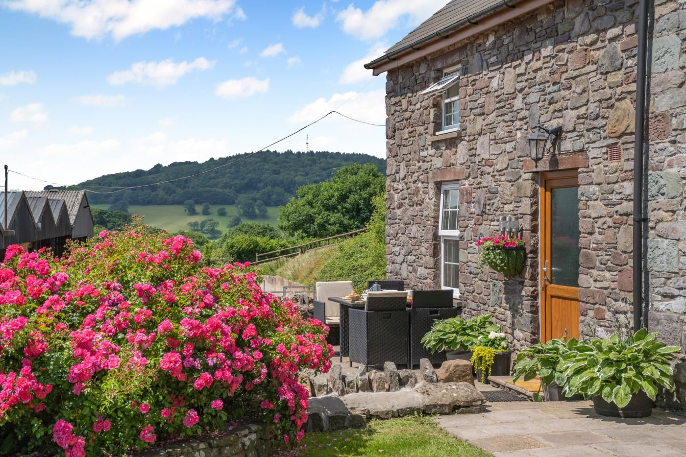 Un bâtiment en pierre avec une porte en bois, une terrasse avec du mobilier d’extérieur et un jardin agrémenté de fleurs roses.