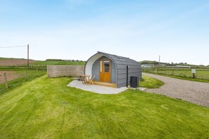 A small, single-story structure with a curved roof, a wooden door, and a concrete patio area with a table and chairs. - 1 Bed in Forfar (oc-t34621) (Forfar)
