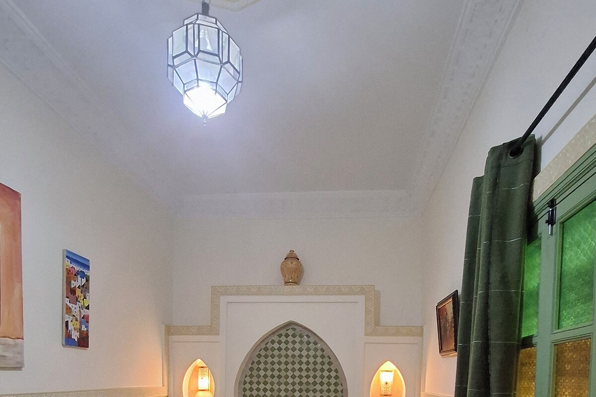 Una habitación con una cama, dos mesitas de noche, una lámpara colgante, piso estampado y una puerta verde.