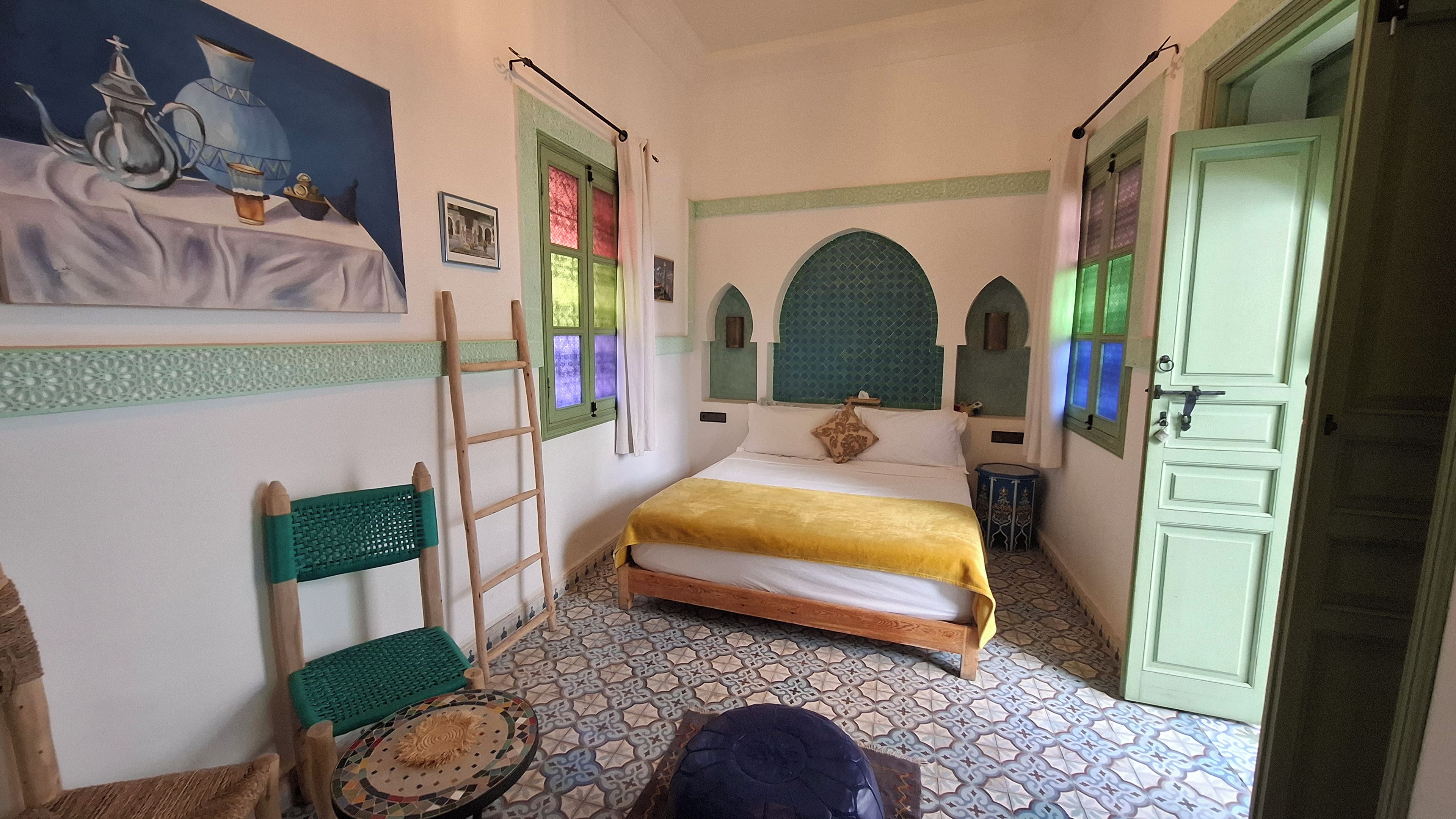 A bedroom with a bed, a ladder, a chair, a small table, and a door.
