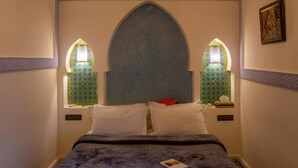 Una habitación con una cama, dos mesitas de noche, un ventanal con marco decorativo y un cuadro colgado en la pared.