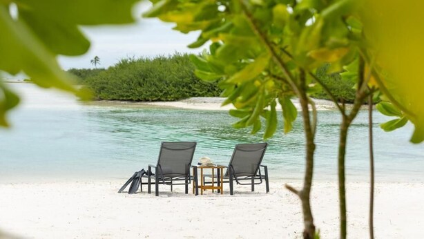 A beach with two chairs and a table, surrounded by clear water and greenery.