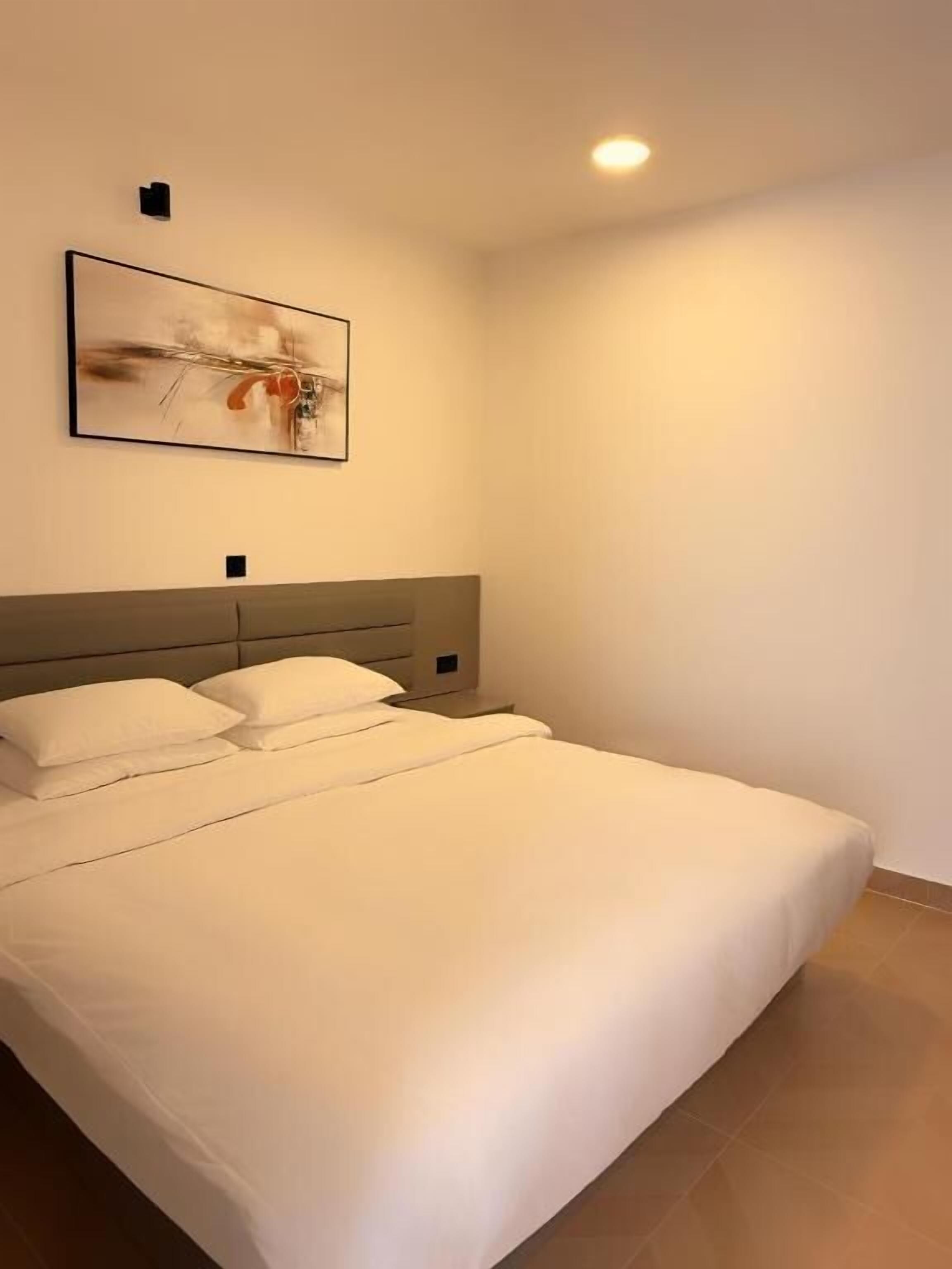 A neatly made bed with a headboard, a wall-mounted light fixture, and a framed abstract painting.