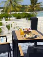 Una mesa con comida, una bebida amarilla y una cesta de fruta, con vista a una playa con palmeras y el océano.