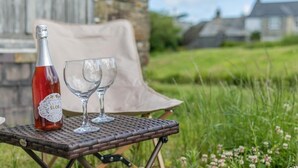 A bottle of Sloe Gin and two glasses on a wicker table outdoors. - 1 Bed in St. Gennys (oc-r34472) (Rosecare)
