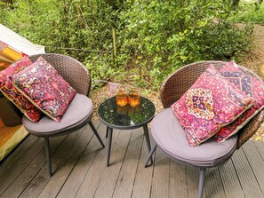 Twee rieten stoelen met kussens en een klein tafeltje met twee kaarsen, op een houten terras met begroeiing op de achtergrond.