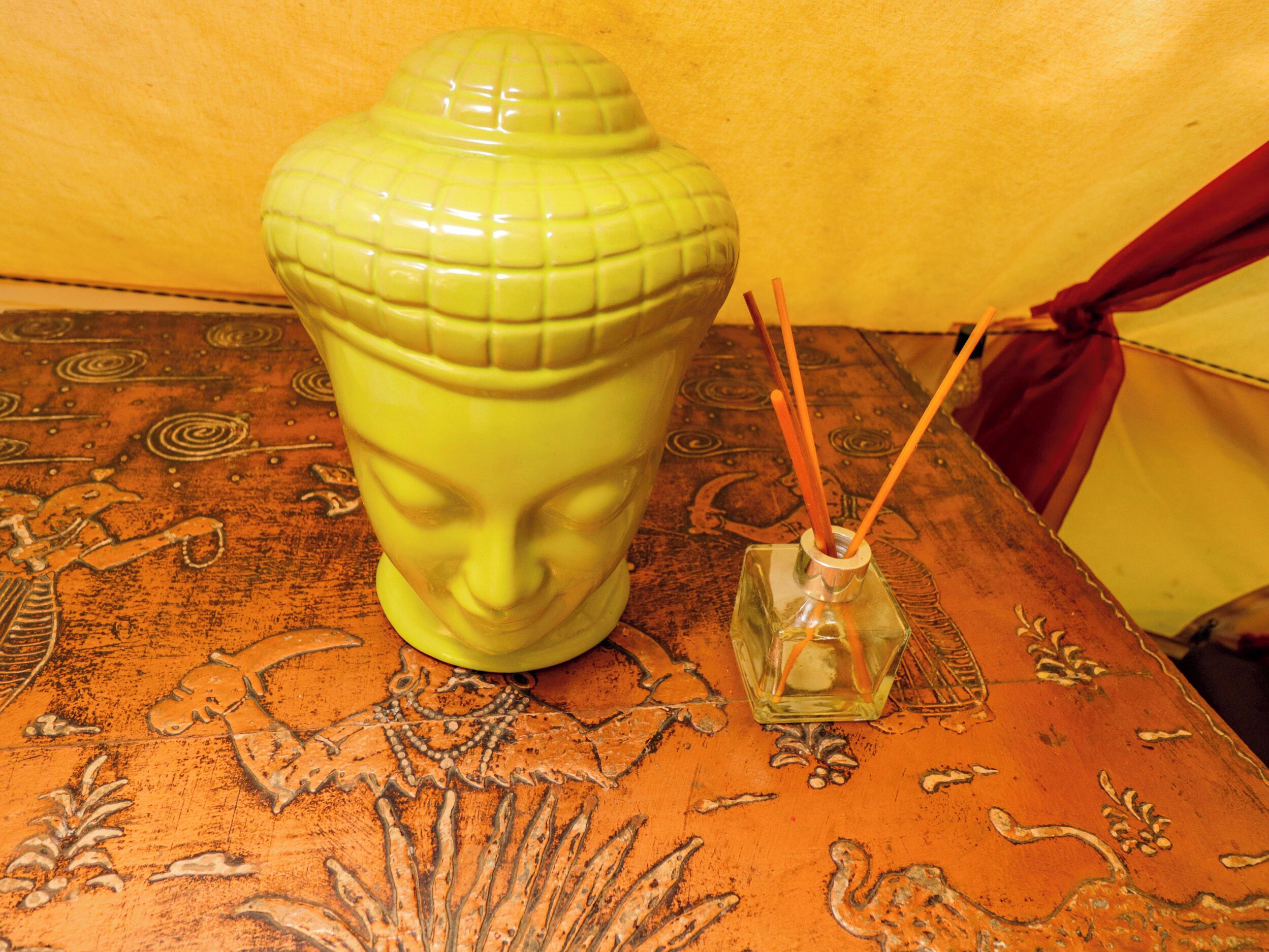 A green vase with a face design and a small glass container with reed sticks on a patterned surface.