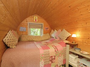 Ein gemütliches Schlafzimmer mit Holzbalkendecke, einem Bett mit floralem Quilt, Kissen mit Mustern, einer Nachttischlampe und einem Fenster mit Blick auf Bäume.