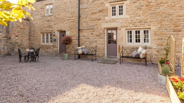 A stone building with a gravel courtyard, a table with chairs, and a bench with cushions. - 3 Bed in Stoney Middleton (oc-c34611) (Stoney Middleton)
