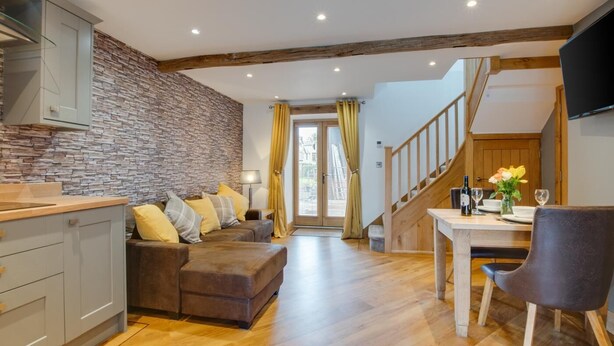 Un espacio habitable moderno con pared de piedra, suelo de madera, un sofá con cojines, una mesa de comedor con sillas y una escalera.