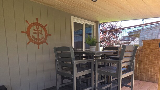 Een overdekt terras met een houten tafel en stoelen, een hondenbak en een aan de muur bevestigd ankerlogo.