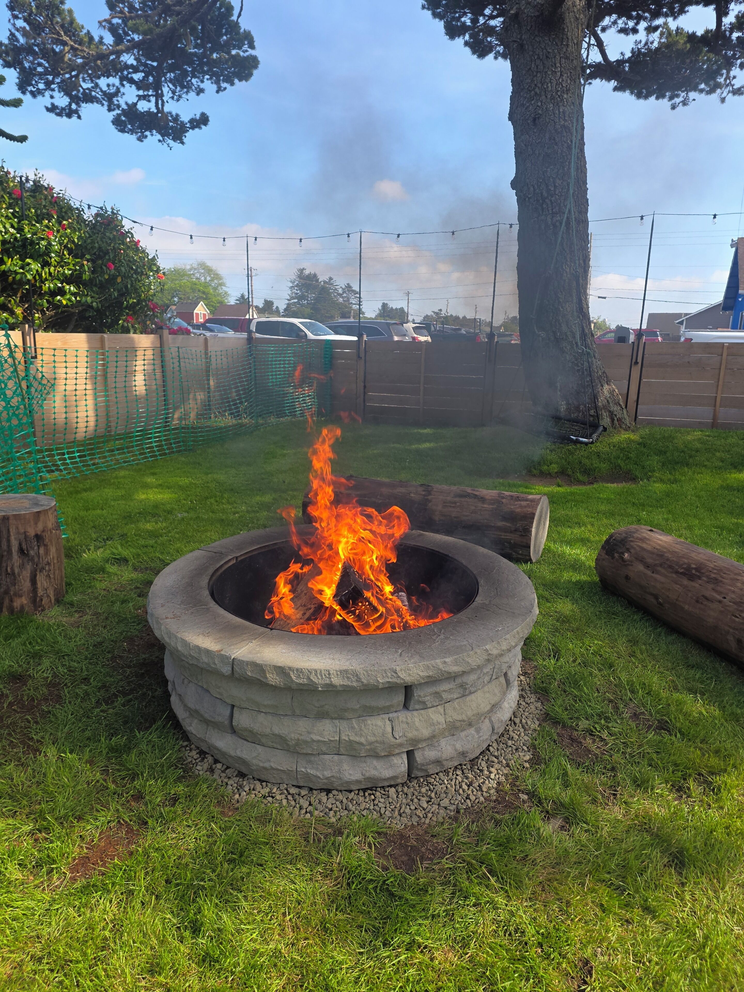 A fire pit with flames, surrounded by a grassy area, a tree, and a fence.