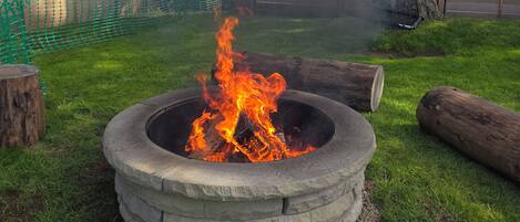 A fire pit with flames, surrounded by a grassy area, a tree, and a fence.