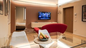 A modern living room with a glass table, a red chair, a bowl of fruit, a television, and a bed in the background. - Apartment 'Flat 1' with Wi-Fi and Air Conditioning (Portese)