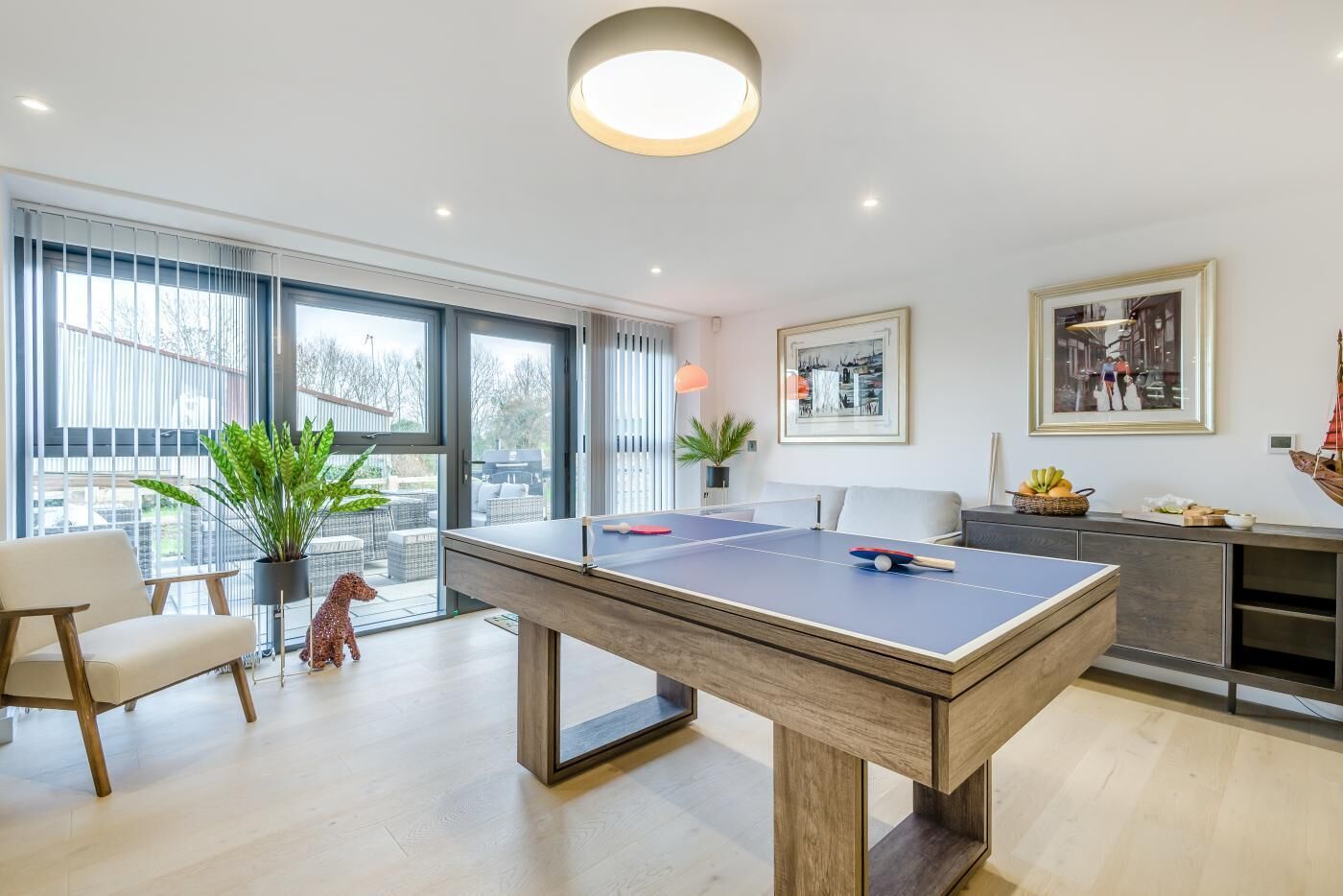 A modern living room with a ping pong table, a sofa, a dining area, and large windows.