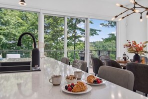 A modern dining area with a large table, chairs, a coffee maker, and a view of trees through large windows. - Bellaria Tremblant - Private villa with SPA (Mont-Tremblant)