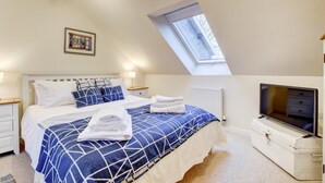 Une chambre avec un plafond en pente, un puits de lumière, un lit recouvert d’une couette à motifs géométriques, une table de chevet avec une lampe et un téléviseur posé sur une commode.