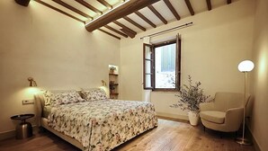 Ein Schlafzimmer mit Holzboden, einem Bett mit floralem Bettzeug, einem Nachttisch, einer Lampe, einem Sessel, einem Fenster mit Rollläden und einem Blumentopf.