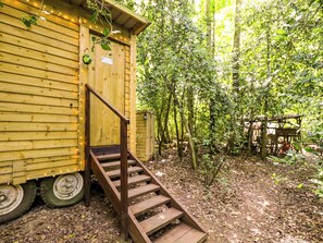 A wooden cabin with a staircase leading to a door, surrounded by trees and a forest floor. - Jungle Bell Tent (Newbury)
