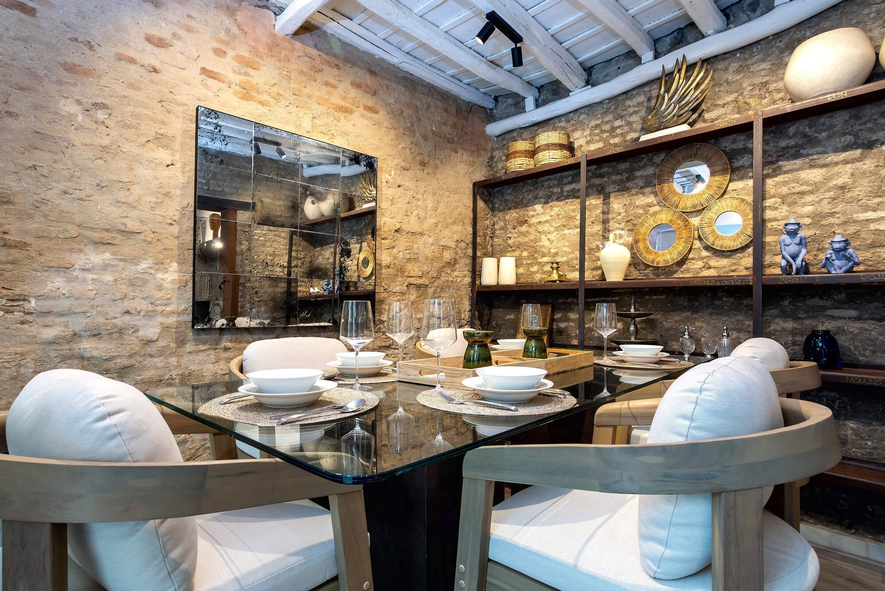 A dining area with a glass table, white cushioned chairs, and a brick wall.