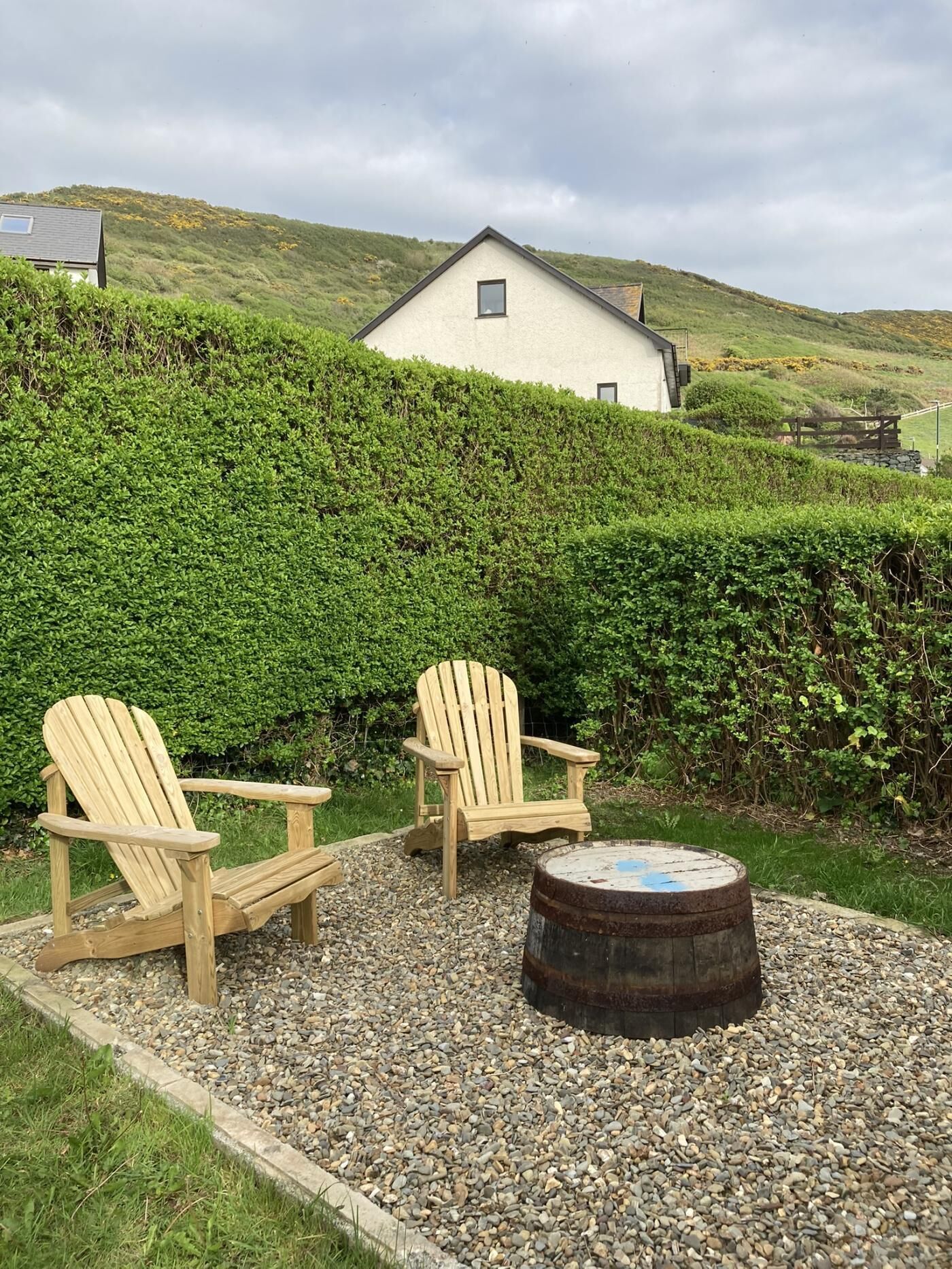 A house with a white exterior, two wooden Adirondack chairs, and a metal barrel on a gravel area.