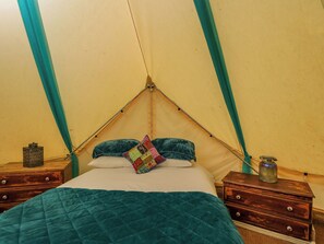 A tent with a bed, pillows, a quilted blanket, a small table with a candle, and a wall-mounted shelf. - The Moroccan Bell Tent (Newbury)