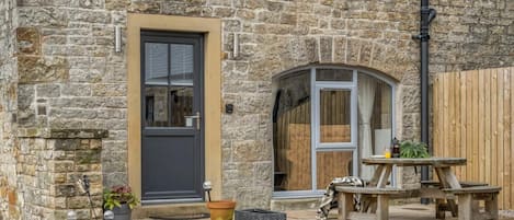 A stone building with a dark door, a small window, and a wooden table with chairs outside.