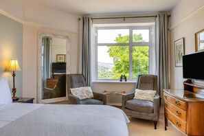Une chambre d’hôtel comprenant un lit, deux fauteuils, une petite table, une commode, un téléviseur et une grande fenêtre donnant sur des arbres.