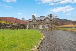 A stone house with a gravel driveway, a stone wall, and a grassy lawn.