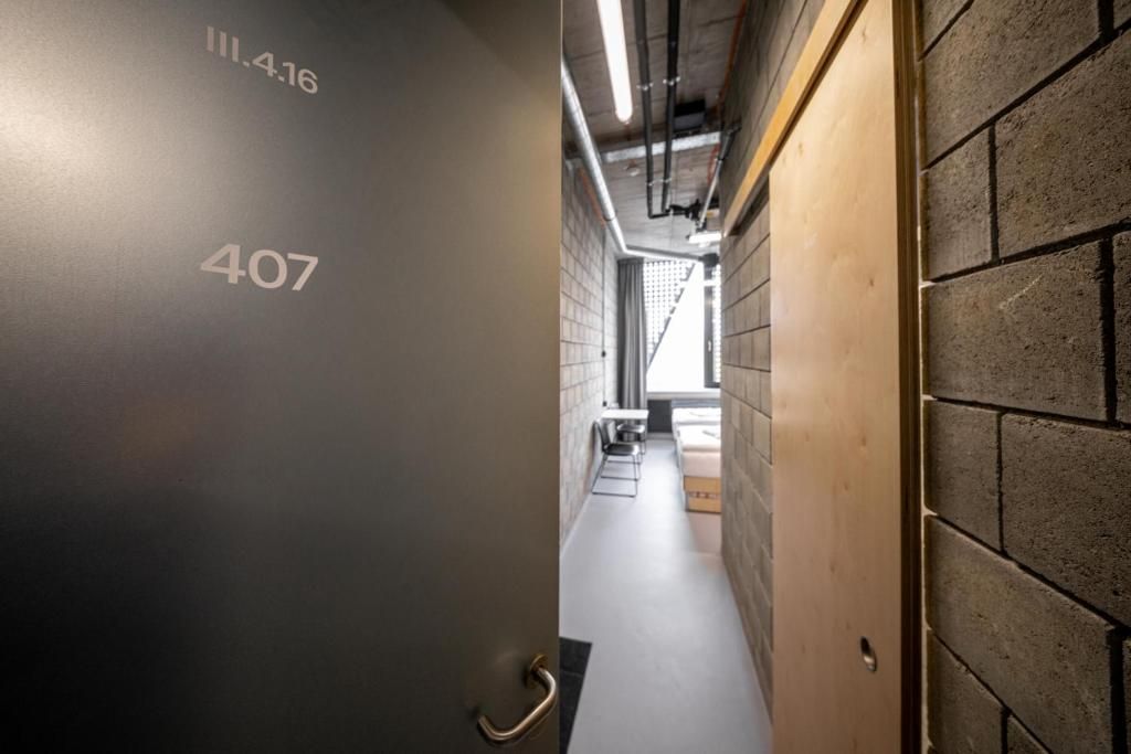 A hallway with a number '407' on the wall, a wooden door on the right, and a view into a room with a bed and a window.