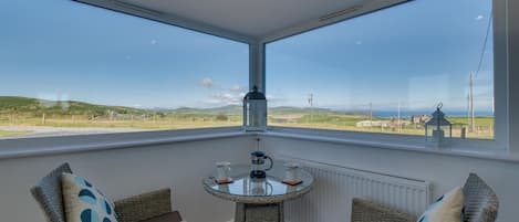 Una habitación con una mesa de vidrio, dos sillas de mimbre y vistas a un paisaje verde a través de amplios ventanales.