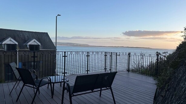 Um deck de madeira com móveis para área externa, guarda-corpo de metal e vista para o mar.