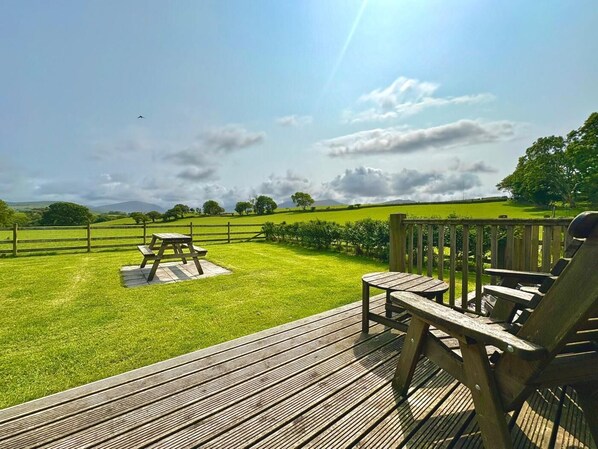 Eine Holzterrasse mit Picknicktischen und -stühlen mit Blick auf ein Rasenfeld, umzäunt von Bäumen.