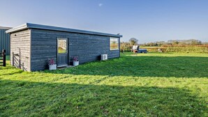 Ein kleines, eingeschossiges Holzgebäude mit Tür und Fenstern, umgeben von einer grünen Rasenfläche und einem Feld im Hintergrund.