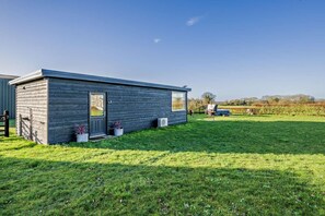 A small, single-story wooden building with a door and windows, surrounded by a grassy lawn and a field in the background. - 1 Bed in Foulsham (oc-j34678) (Foulsham)