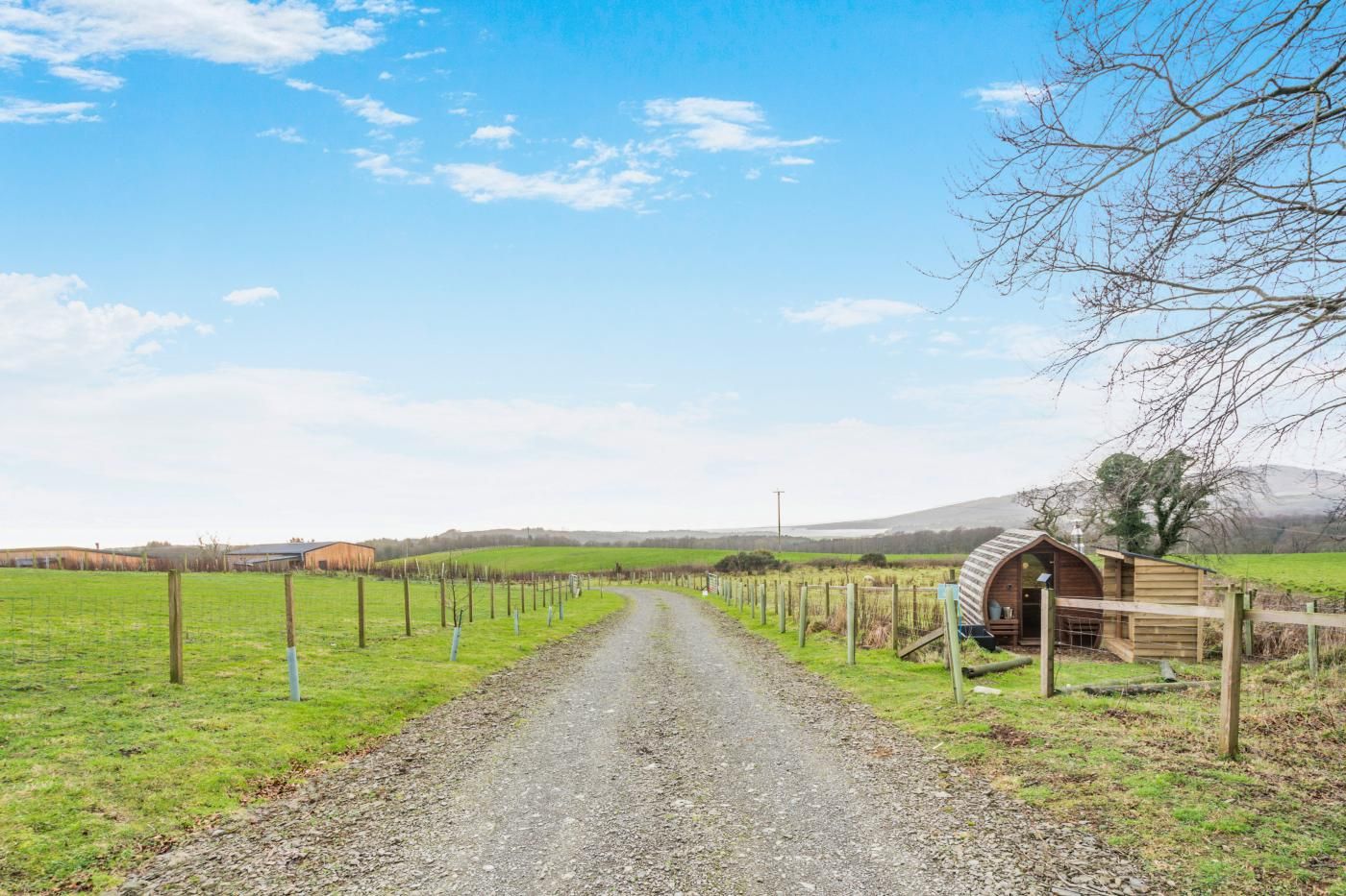 A rural road leading to a wooden structure with a curved roof, surrounded by a fence and fields.
