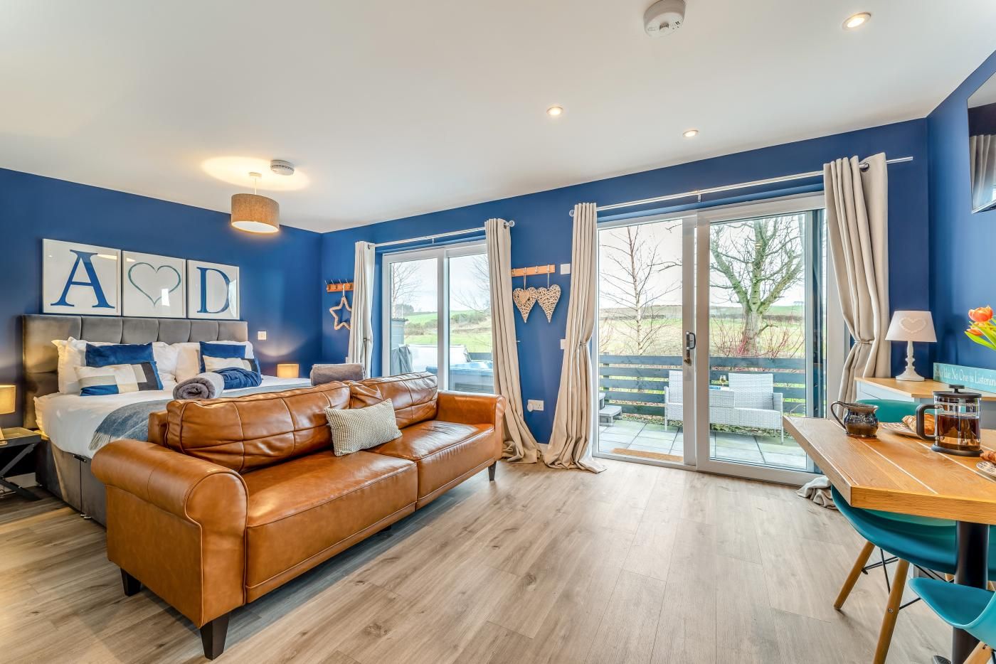 A room with a bed, a leather sofa, a dining table, and a sliding glass door leading to an outdoor area.
