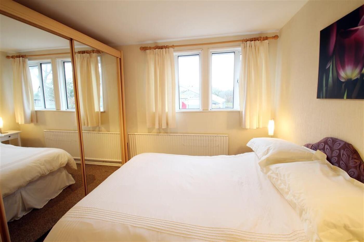A hotel room with a large bed, a mirror, two windows with curtains, a radiator, and a painting on the wall.