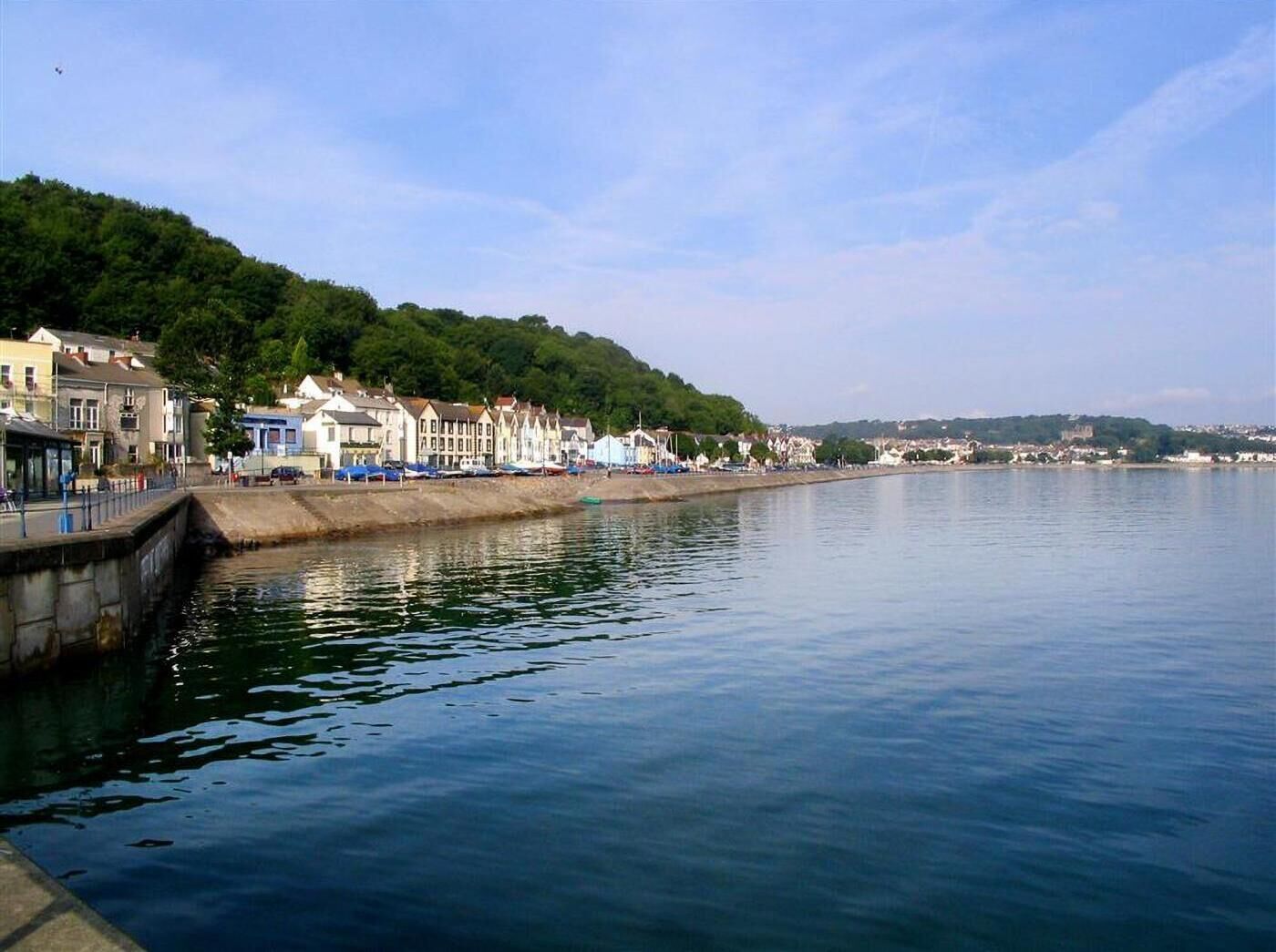 A waterfront view with buildings along the shore, a calm body of water, and a hilly background.