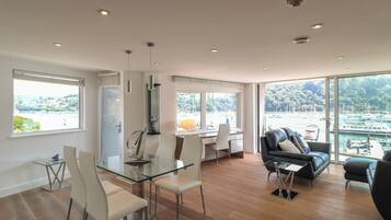 A modern living room with a glass dining table, white chairs, a leather sofa, and large windows offering a view of a marina.