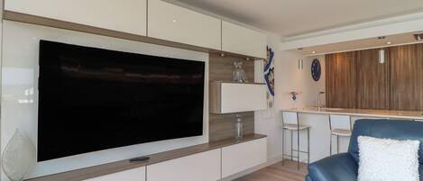 Una sala moderna con un televisor de pantalla plana grande, armarios empotrados, un sofá de cuero azul y una zona de cocina con armarios de madera y un reloj.