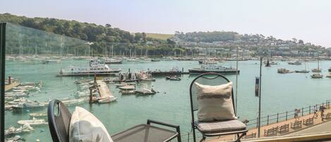 A balcony with two chairs and a small table, overlooking a marina with boats and a distant town.
