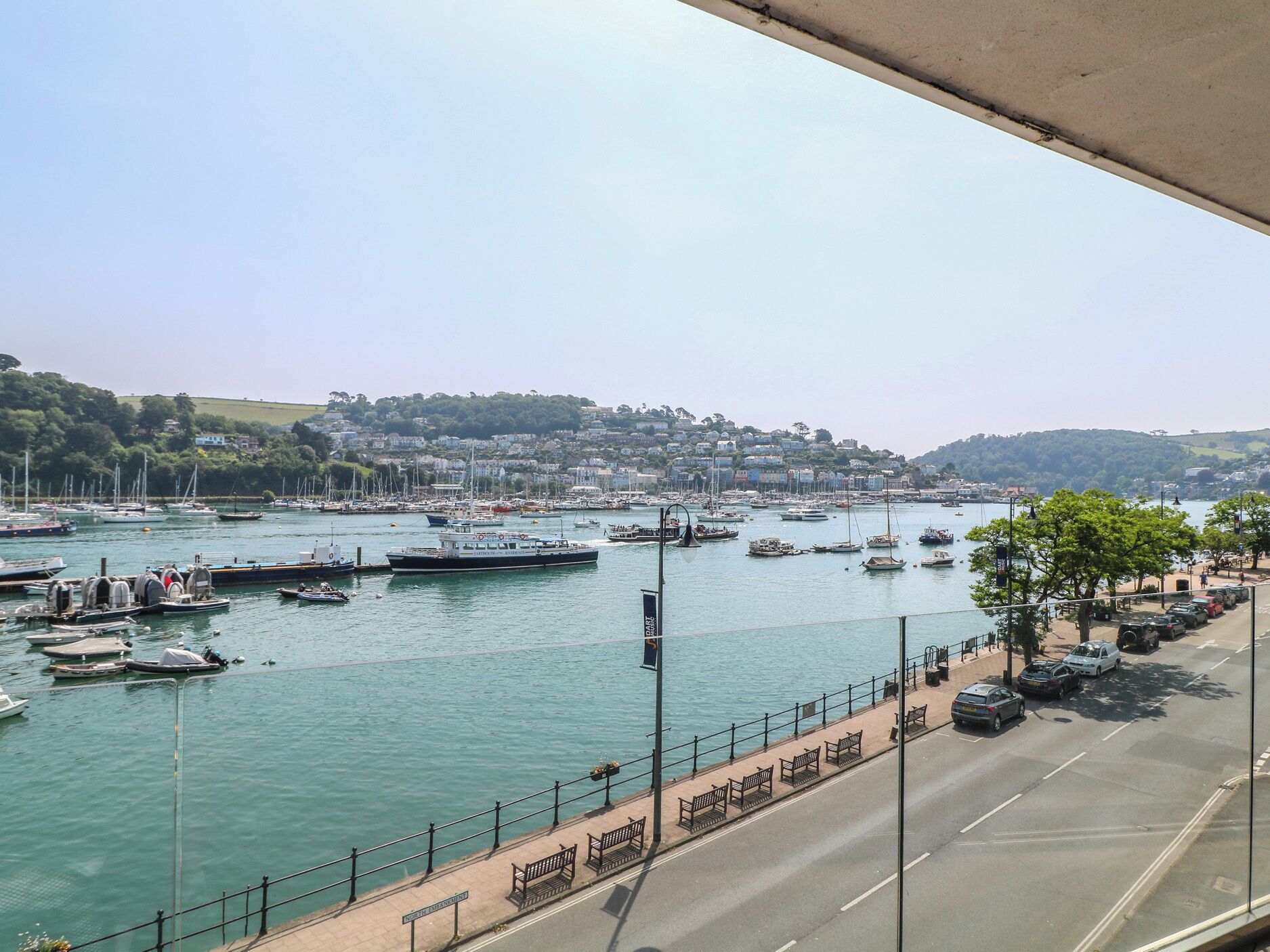 A marina with boats docked, a road with parked cars, and a pedestrian walkway.