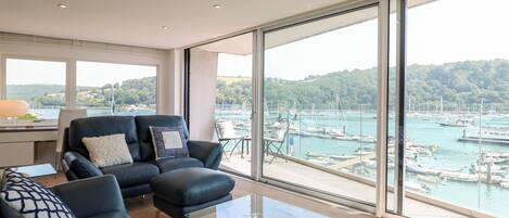 Un salón moderno con una mesa de centro de vidrio, un sofá de cuero negro y vista a un puerto deportivo.