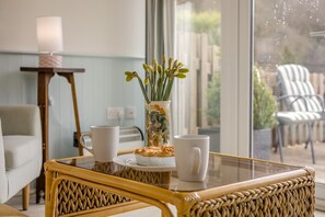 A room with a wicker chair, a glass table with a pastry, two white mugs, and a vase with flowers. - 1 Bed in Caswell, Gower (oc-hh375) (Newton)