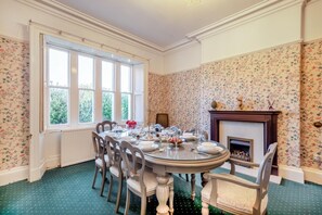 Une salle à manger avec une table ronde dressée pour six personnes, flanquée de deux fenêtres, d’une cheminée et d’un papier peint à motifs floraux.