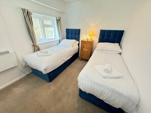 A hotel room with two beds, a window with curtains, a bedside table with a lamp, and a radiator. - 1 Bed in Caswell (oc-hh901) (Bishopston)