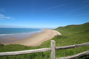 A coastal landscape with a sandy beach, grassy hills, and a wooden fence. - 4 Bed in Gower (oc-hh151) (Llangennith)